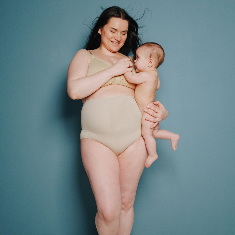 postpartum woman in maternity bra and underwear standing up holding baby on hip