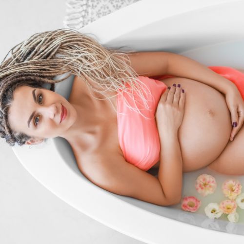 pregnant woman sitting in a bath with flowers wearing a scarf over her chest and looking up smiling