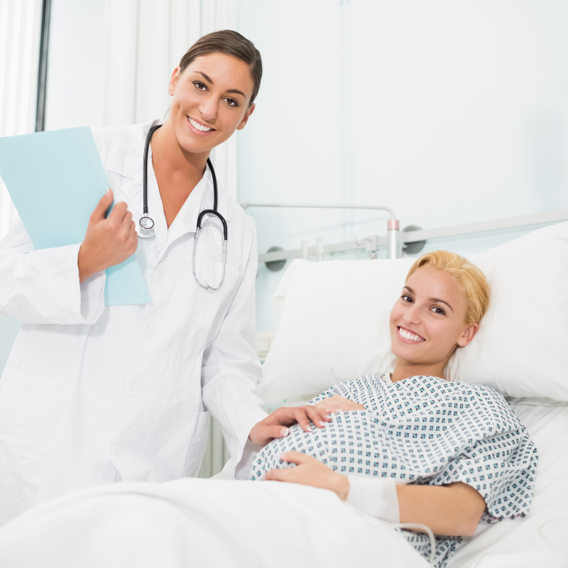 female doctor with stethoscope smiling and holding clipboard, pregnant woman in bed smiling and holding belly